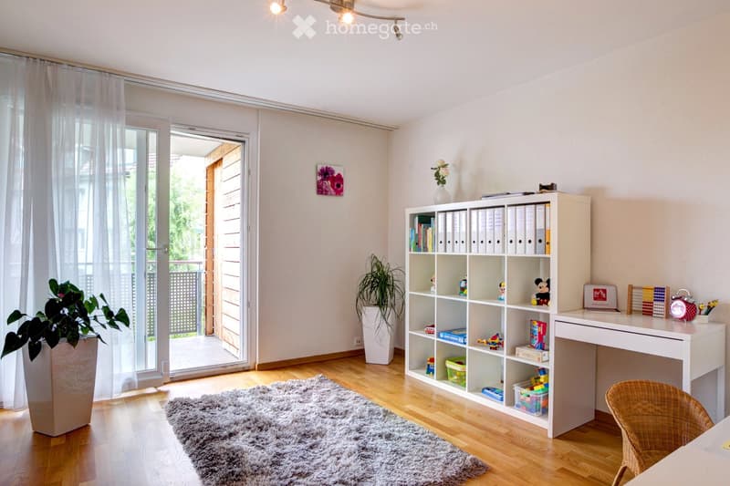 Bright, modern room with wooden flooring, large window with sheer curtains, white desk, wicker chair, white shelving unit filled with books and toys, potted plants, and a fluffy grey rug.