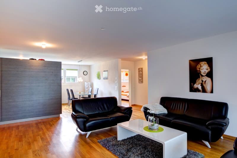 Modern living room with dark leather sofas, white coffee table, grey rug, and a dining area visible in the background.