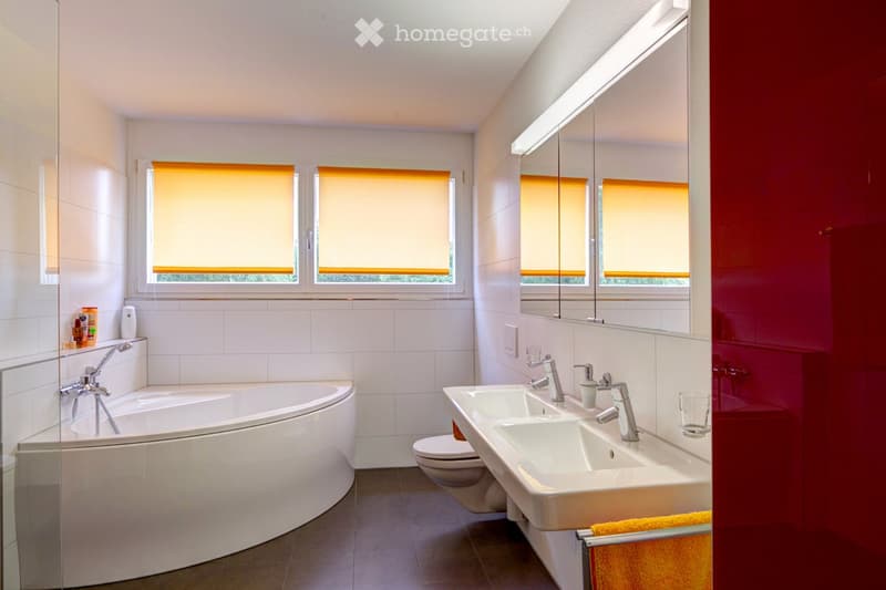Modern bathroom with a large bathtub, double sink, and a large mirror. The room features white tiles, a glass shower enclosure, and a red accent wall.