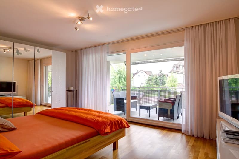 Bright bedroom with wooden flooring, large sliding glass doors leading to a balcony, a bed with orange bedding, a TV, and a mirror.