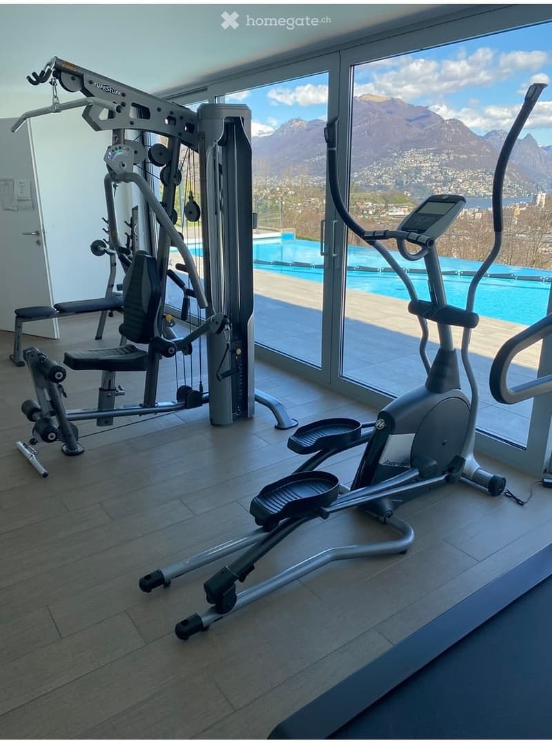 Modern fitness area with elliptical trainer, multi-gym, and bench, large windows offering a scenic mountain view and a swimming pool.