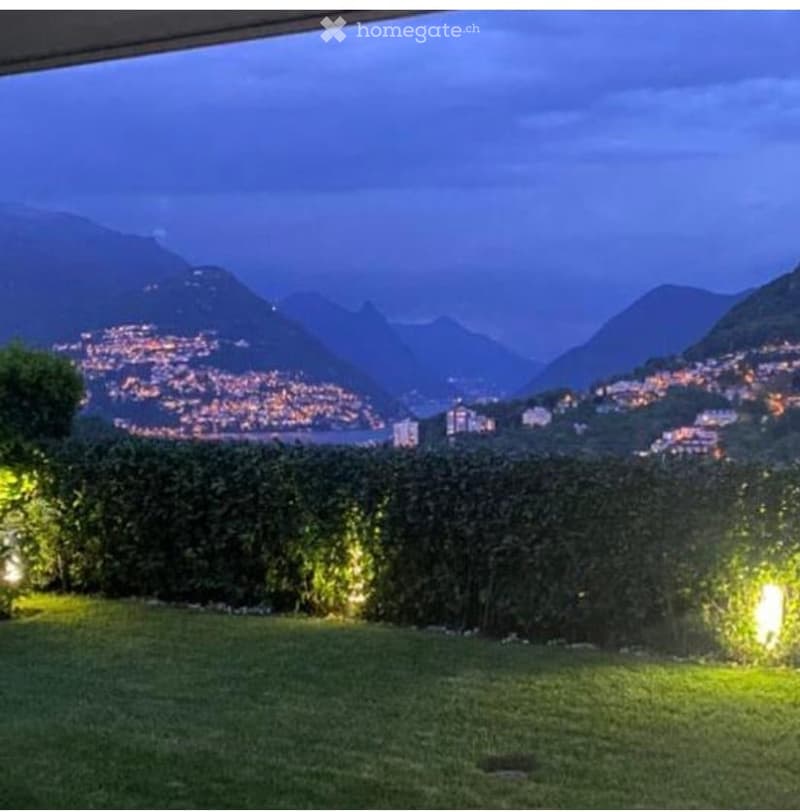 Evening view of a garden with well-lit hedges and a cityscape in the background, featuring mountains and illuminated buildings.