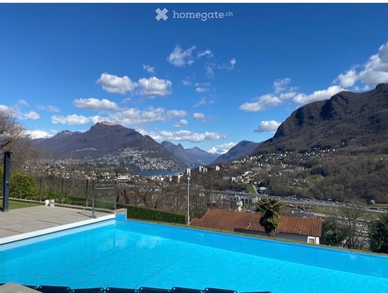Outdoor area with a swimming pool, surrounded by a wooden deck, overlooking a scenic mountain and lake view.