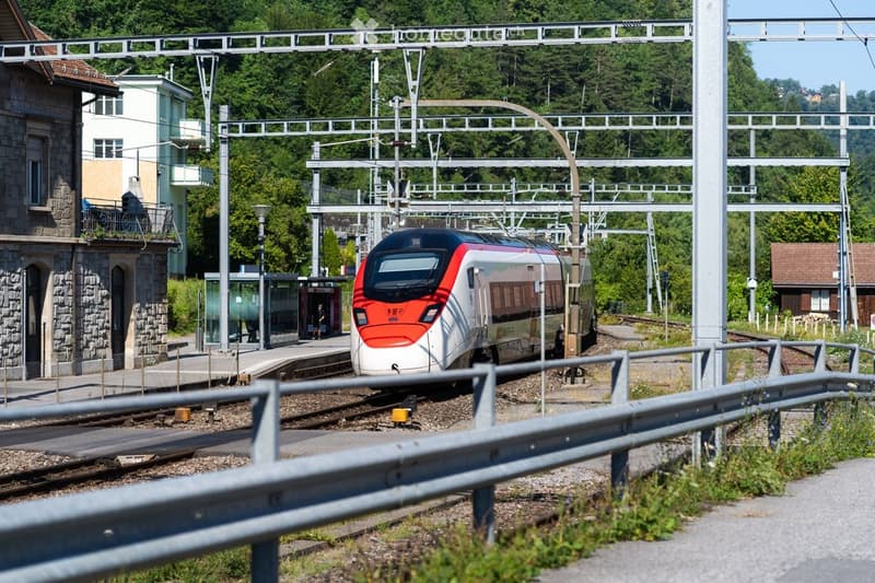 A modern train station with a sleek red and white train arriving at the platform, surrounded by greenery and a stone building.