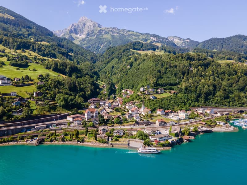A picturesque village nestled in a lush green valley, surrounded by mountains and a turquoise lake. The village features traditional houses, a church, a marina with boats, and a ferry crossing the lake.