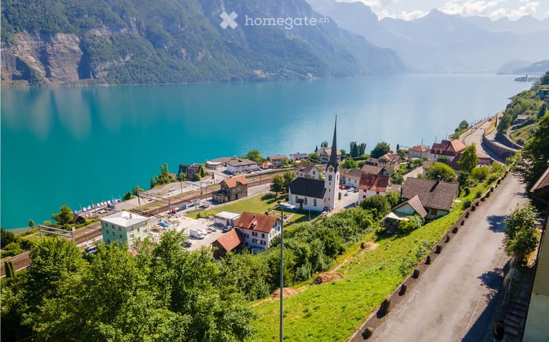 Aerial view of a small village by a lake, surrounded by mountains, with houses, a church, and a road running alongside the water.