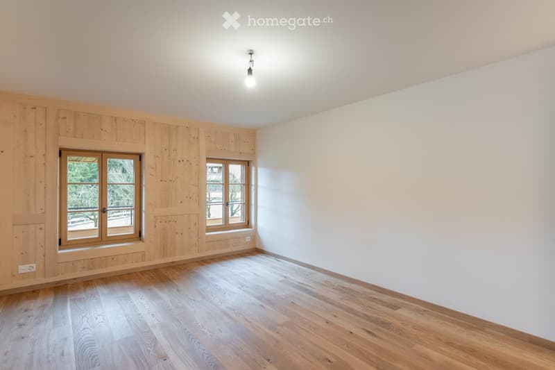 Empty room with wooden flooring, white walls, and large windows with wooden frames. The room has a simple, clean design with no furniture or decorations.