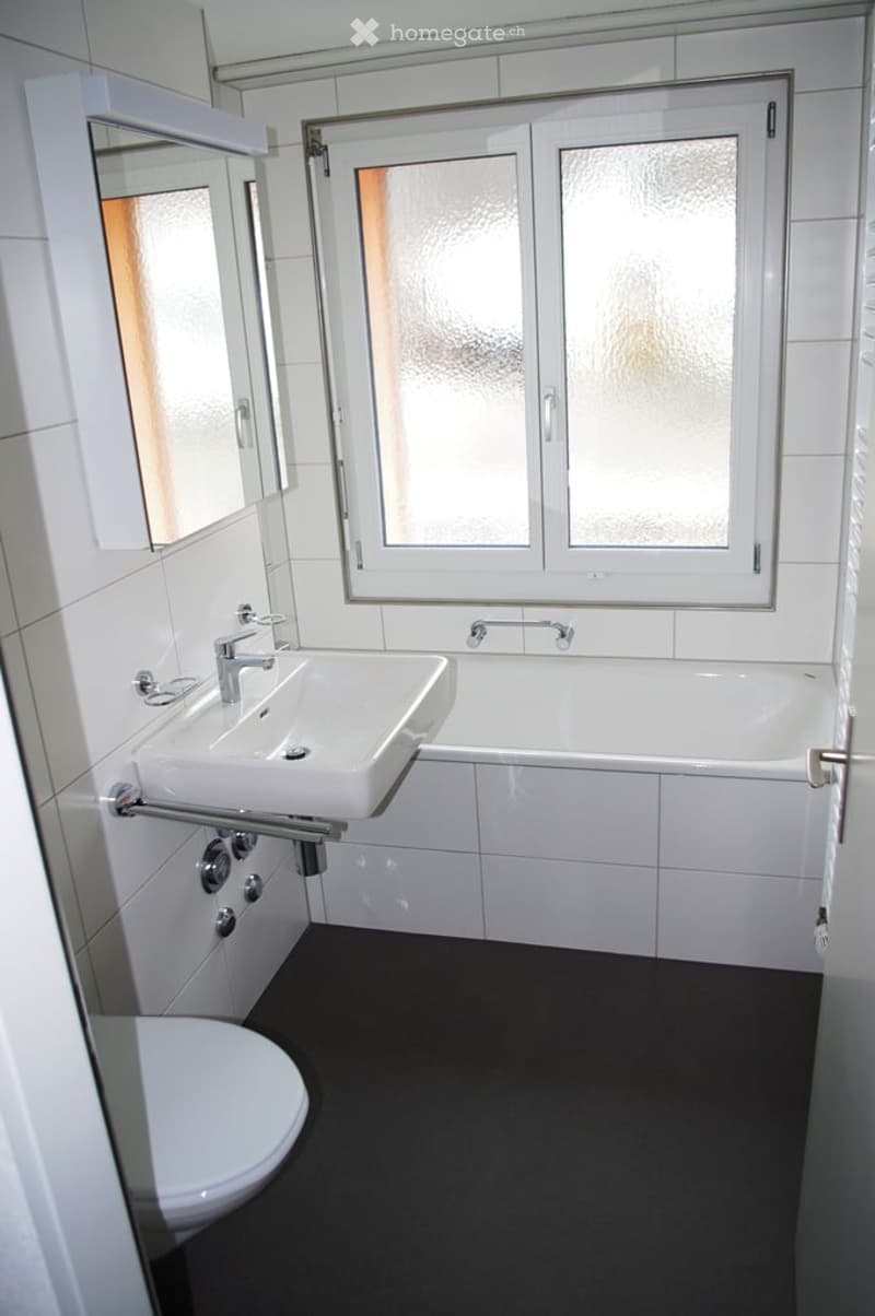 Modern bathroom with white tiles, a sink, bathtub, toilet, and a large window with frosted glass. The floor is dark grey.
