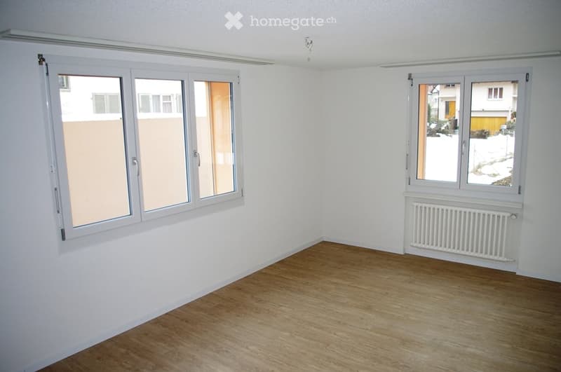 Empty room with wooden flooring, white walls, and large windows with white frames. A radiator is visible on one wall.