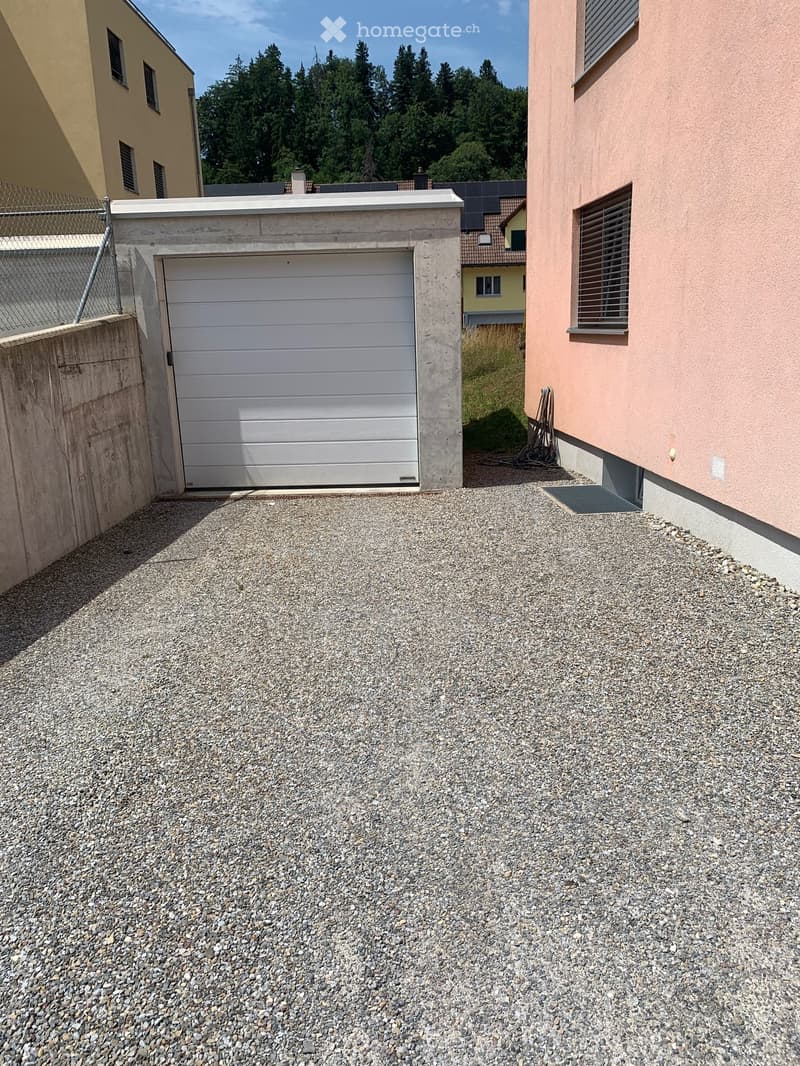 Gravel driveway leading to a single-car garage with a white door, adjacent to a pink house with windows and a small fence.