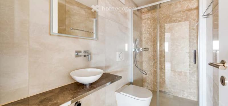 Modern bathroom with a white sink, mirror, toilet, and glass-enclosed shower. Light-colored tiles and sleek fixtures.
