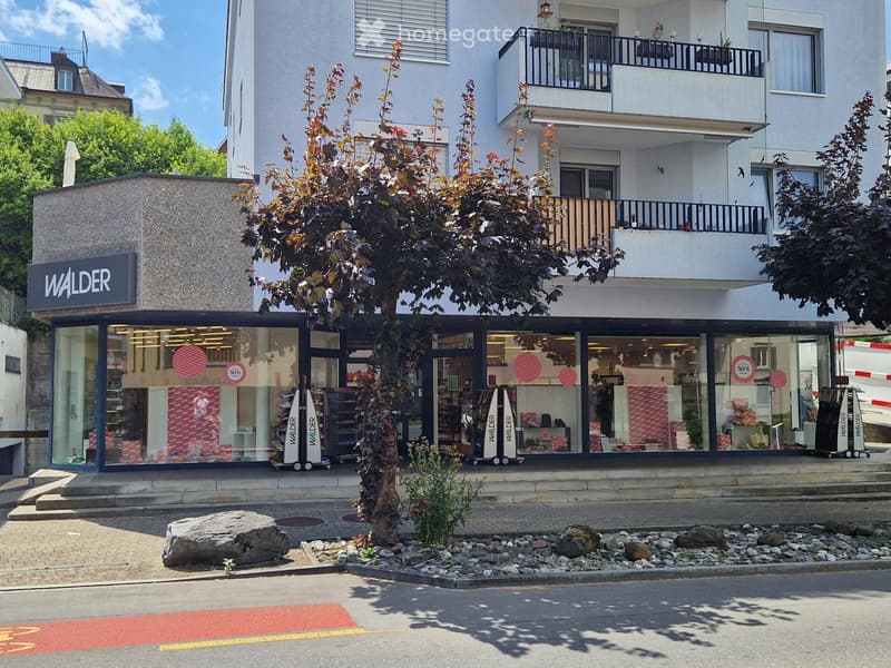 A modern commercial building with a storefront labeled "WALDER" and a balcony above. The storefront has large glass windows displaying items and promotional signs. There is a small landscaped area with rocks and a tree in front of the building.