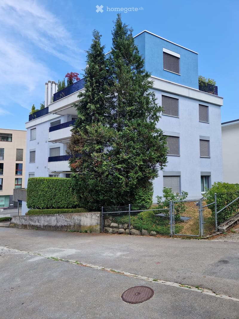 Modern multi-story building with white and blue exterior, surrounded by greenery and a fence.