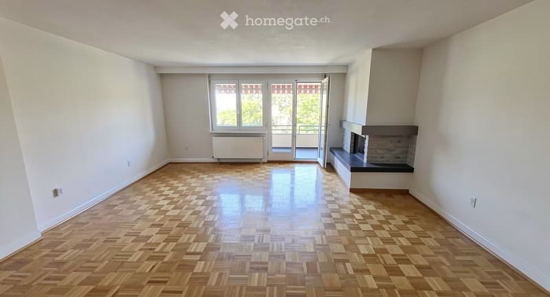 Empty room with wooden parquet flooring, white walls, fireplace, and large window with balcony access.