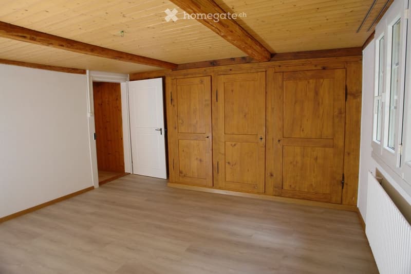 Empty room with wooden flooring, white walls, exposed wooden beams on the ceiling, and built-in wooden cabinetry along one wall. A white door leads to another room.