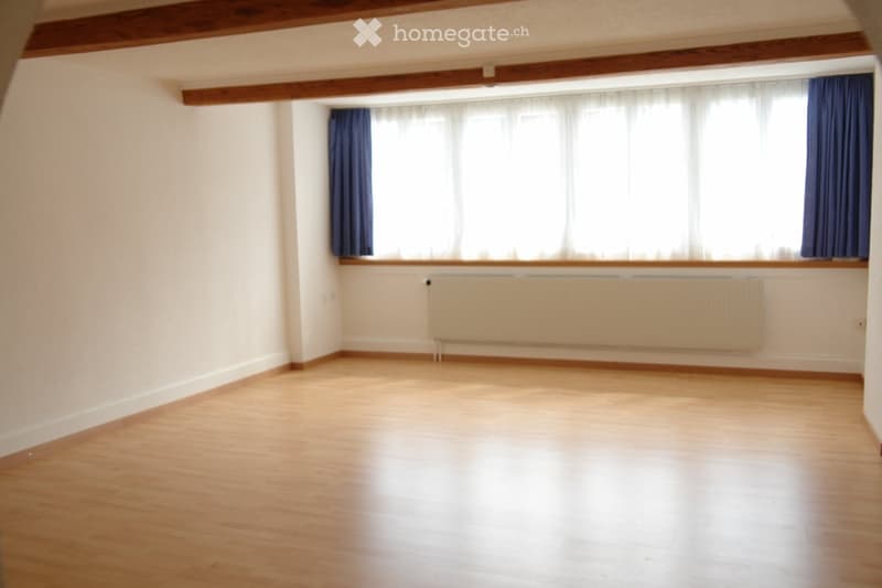 Empty room with wooden flooring, white walls, large window with blue curtains, and a radiator below the window.
