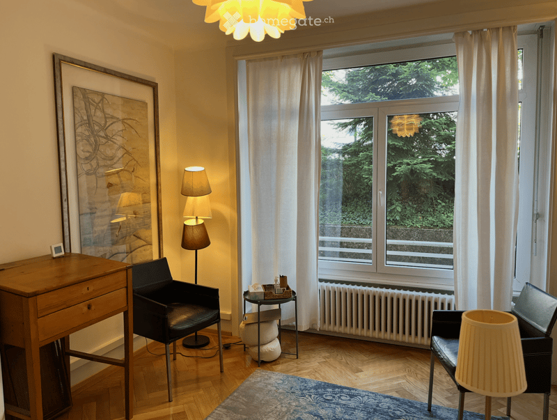 A cozy living room with wooden flooring, large windows with white curtains, a black leather armchair, a small round table with a lamp, a wooden cabinet, and a large picture frame on the wall.