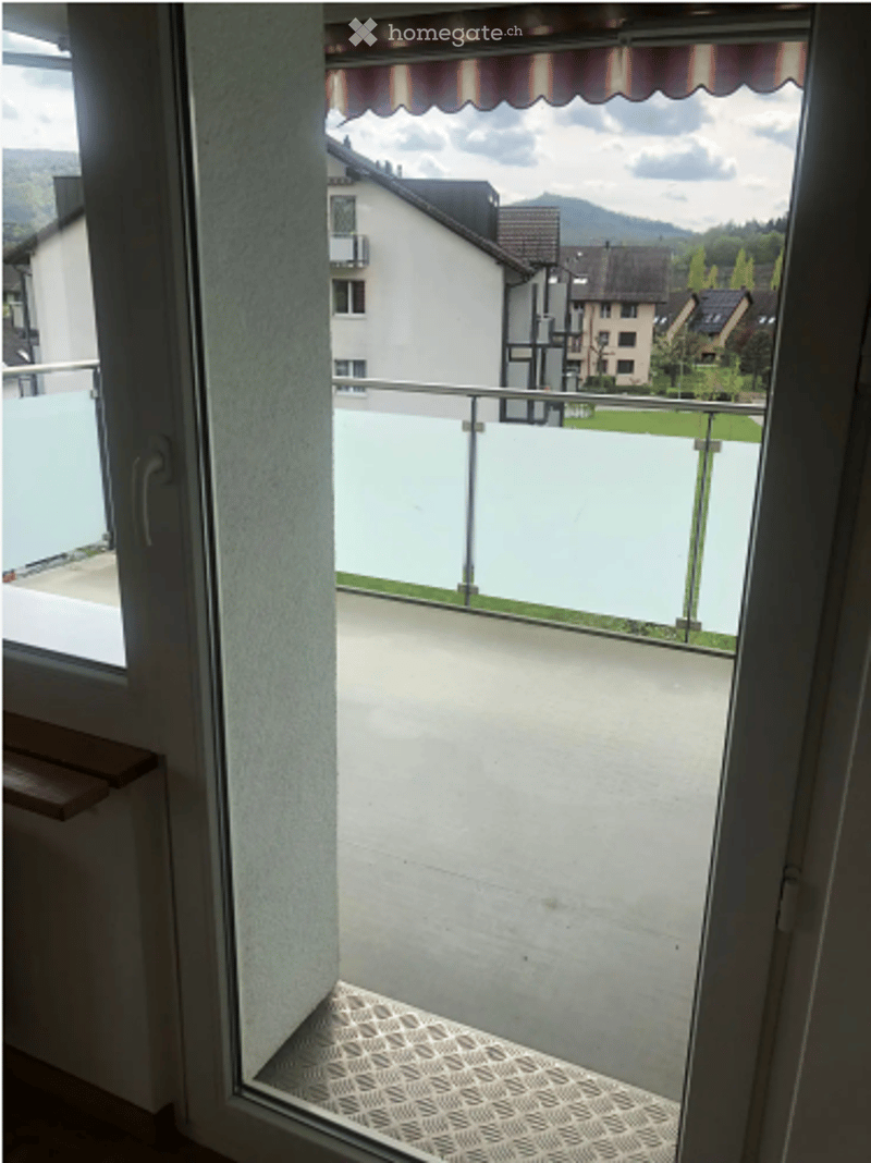 Balcony with glass railing, wooden bench, and view of neighboring houses and mountains.