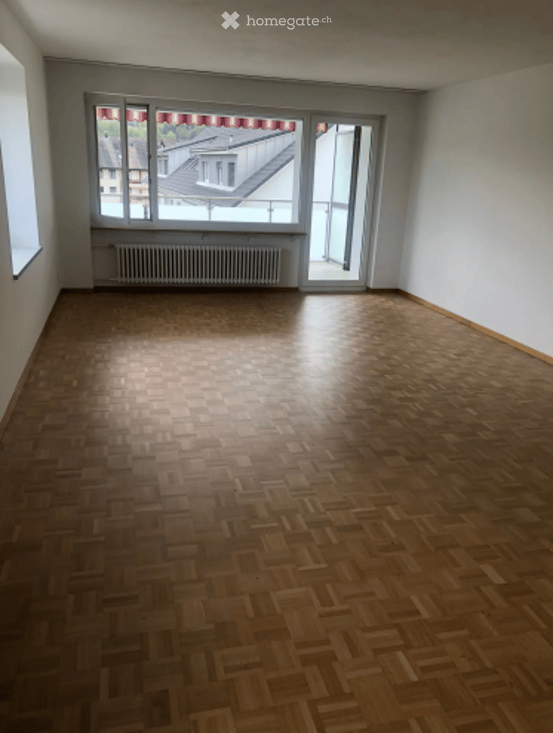 Empty room with wooden parquet flooring, large windows, radiator, and a view of a rooftop.