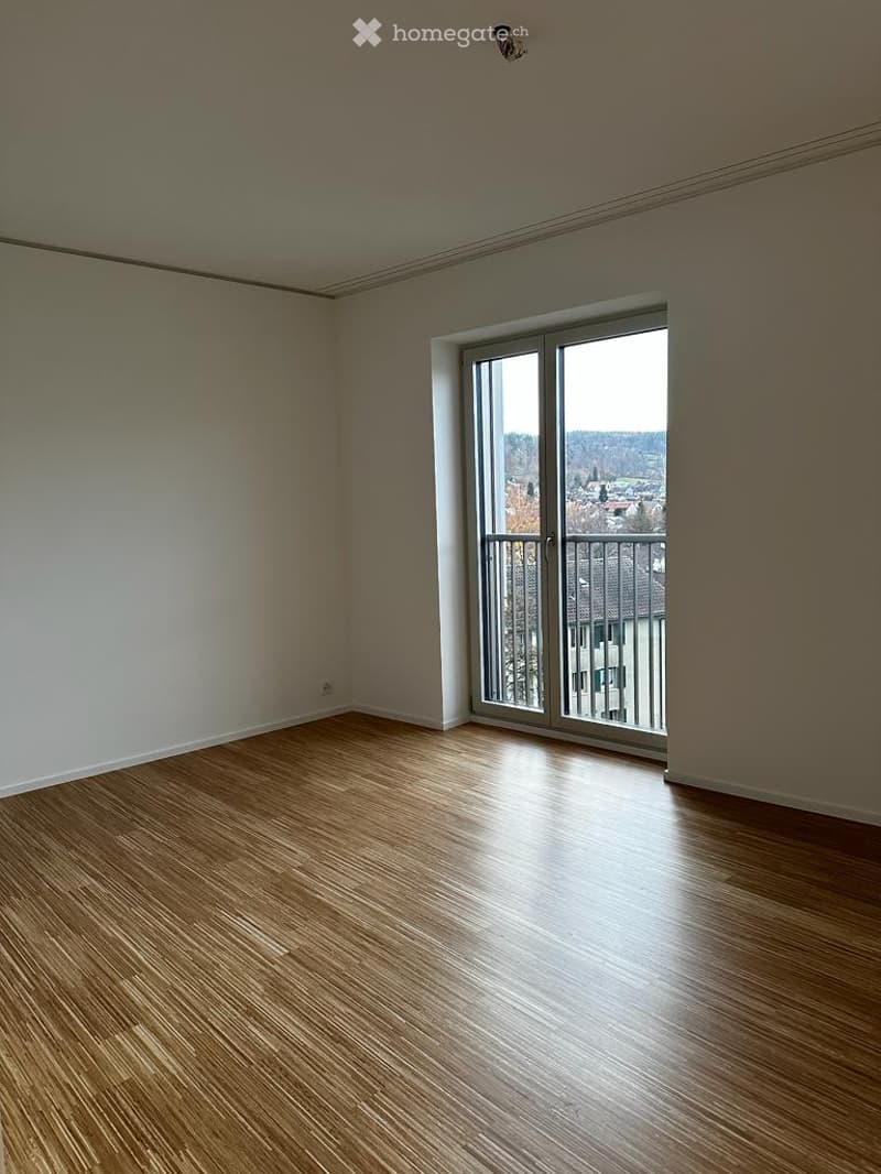 Empty room with wooden flooring, large sliding glass door leading to a balcony with a view of the city and surrounding landscape.
