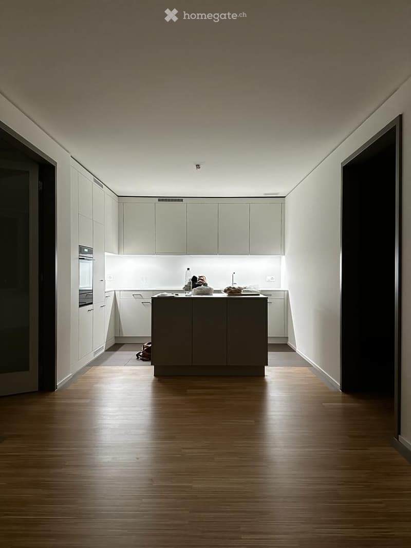 Modern kitchen with white cabinets, a central island, and wooden flooring. Visible items include a bottle and an oven.