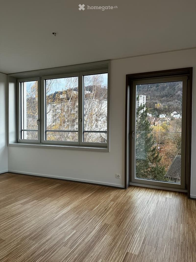 Empty room with wooden flooring, large windows, and a sliding glass door leading to a view of trees and buildings.
