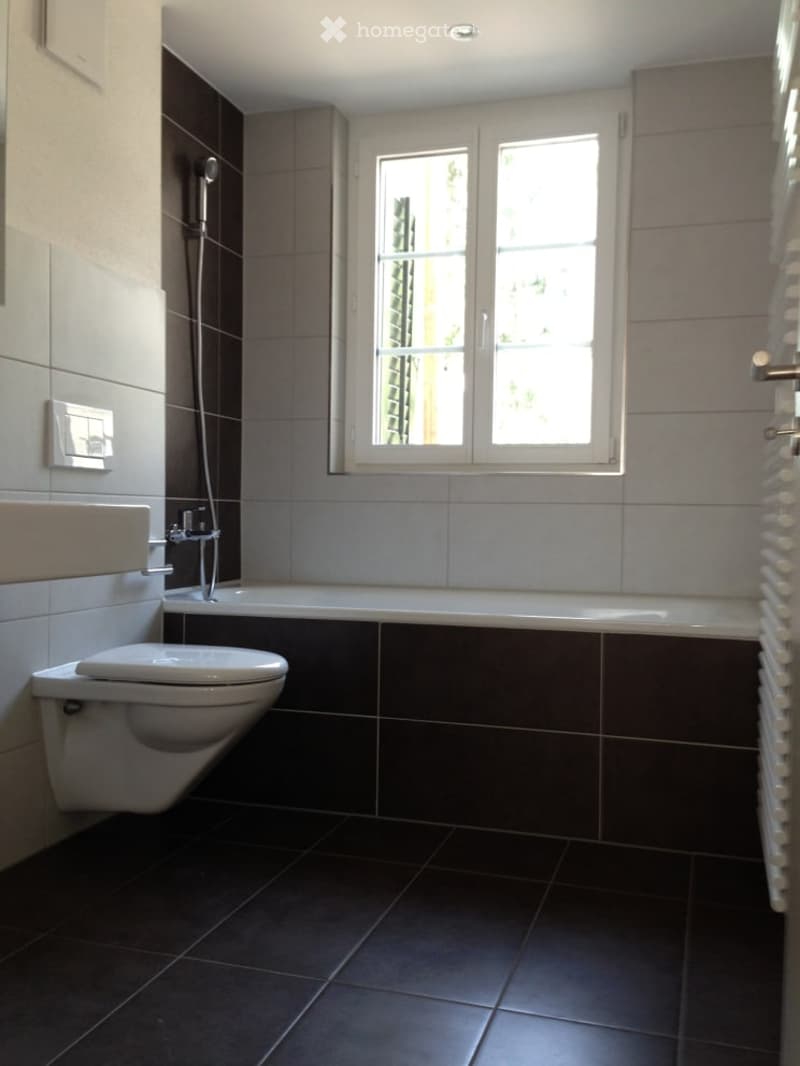 Modern bathroom with dark tiled walls and floor, a toilet, a bathtub, and a showerhead. The room features a window with blinds and a sleek, contemporary design.