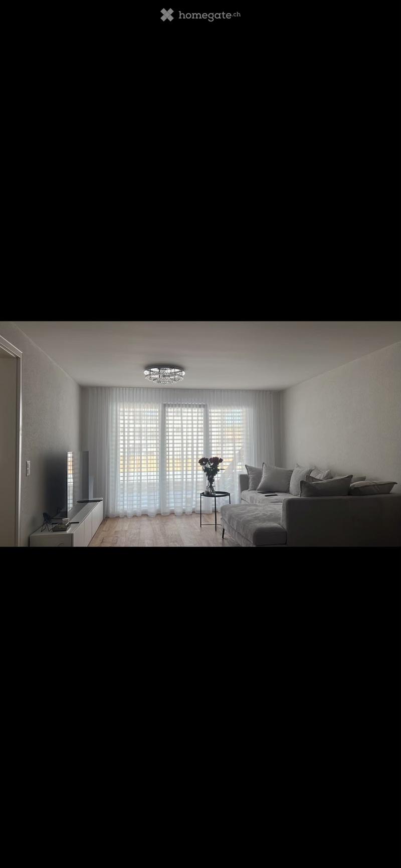 Modern living room with a grey sofa, coffee table, flower vase, and a television on a stand. The room features large windows with sheer curtains and a chandelier on the ceiling.