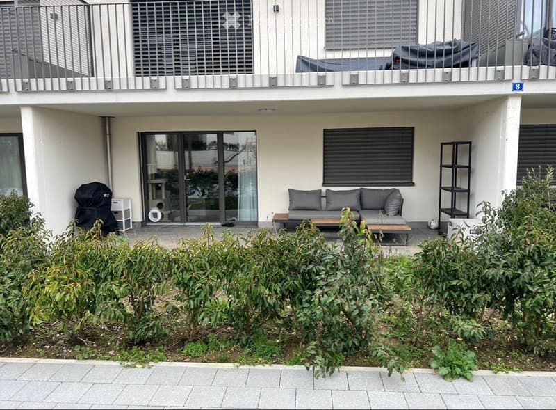 Outdoor patio area with a grey sofa, coffee table, shelving unit, and covered grill. The area is surrounded by greenery and has access to a balcony above.
