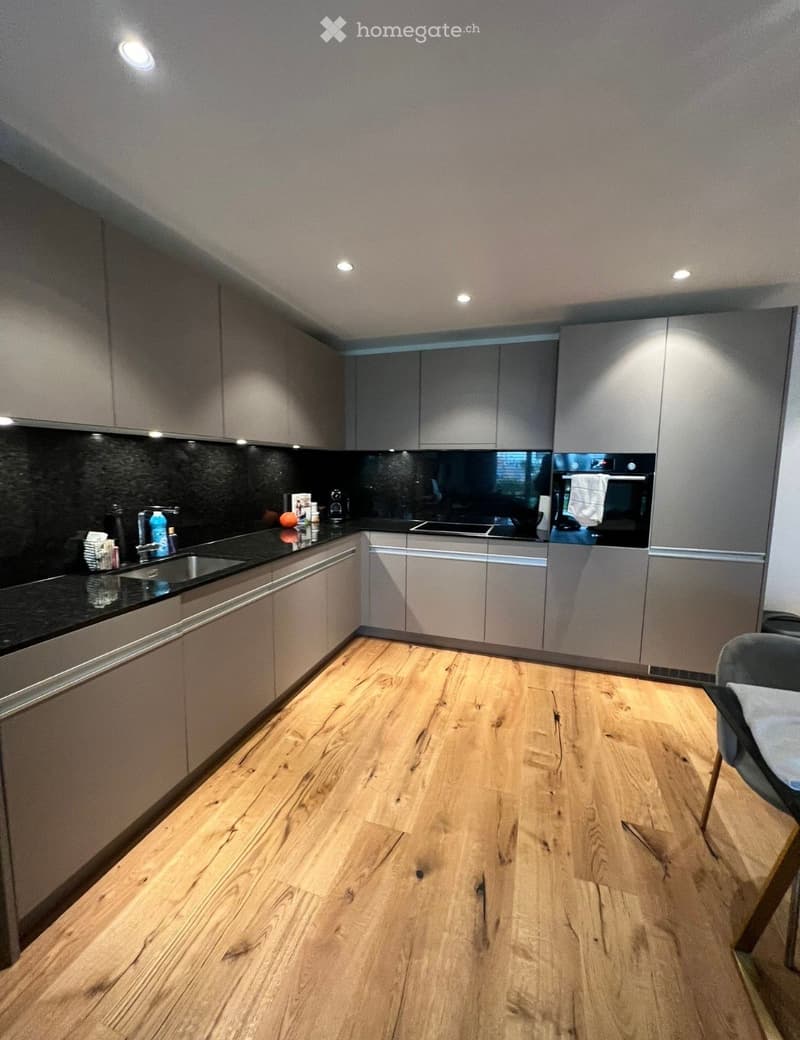 Modern kitchen with light grey cabinets, wooden flooring, and a black countertop. Features a sink, oven, and various small items on the counter.
