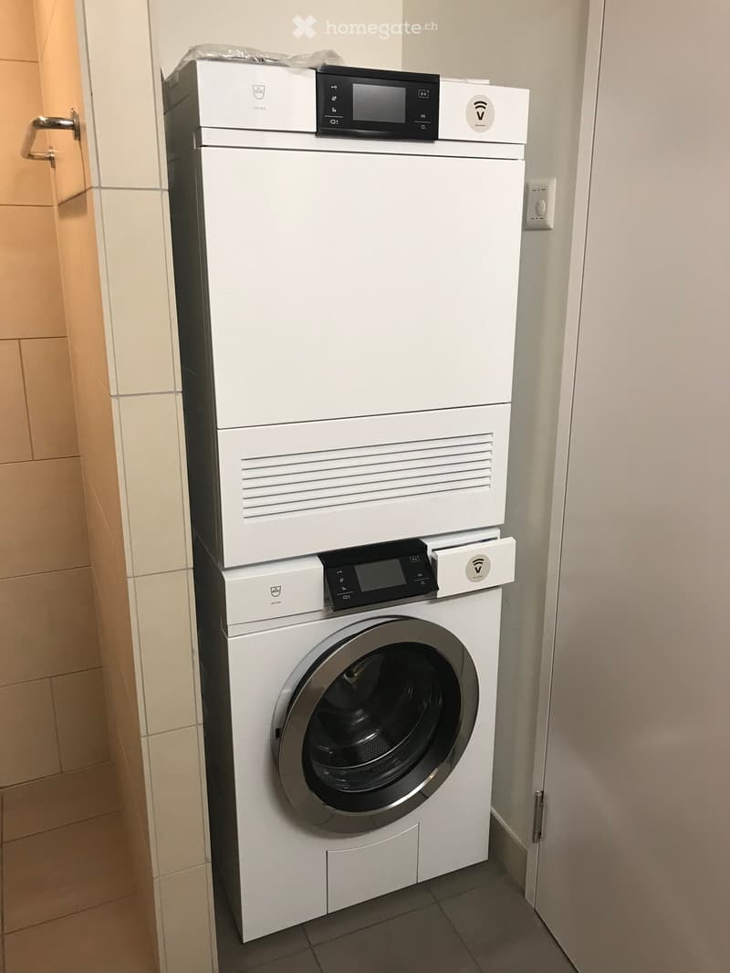 A compact, stacked washing machine and dryer unit in a tiled bathroom area.