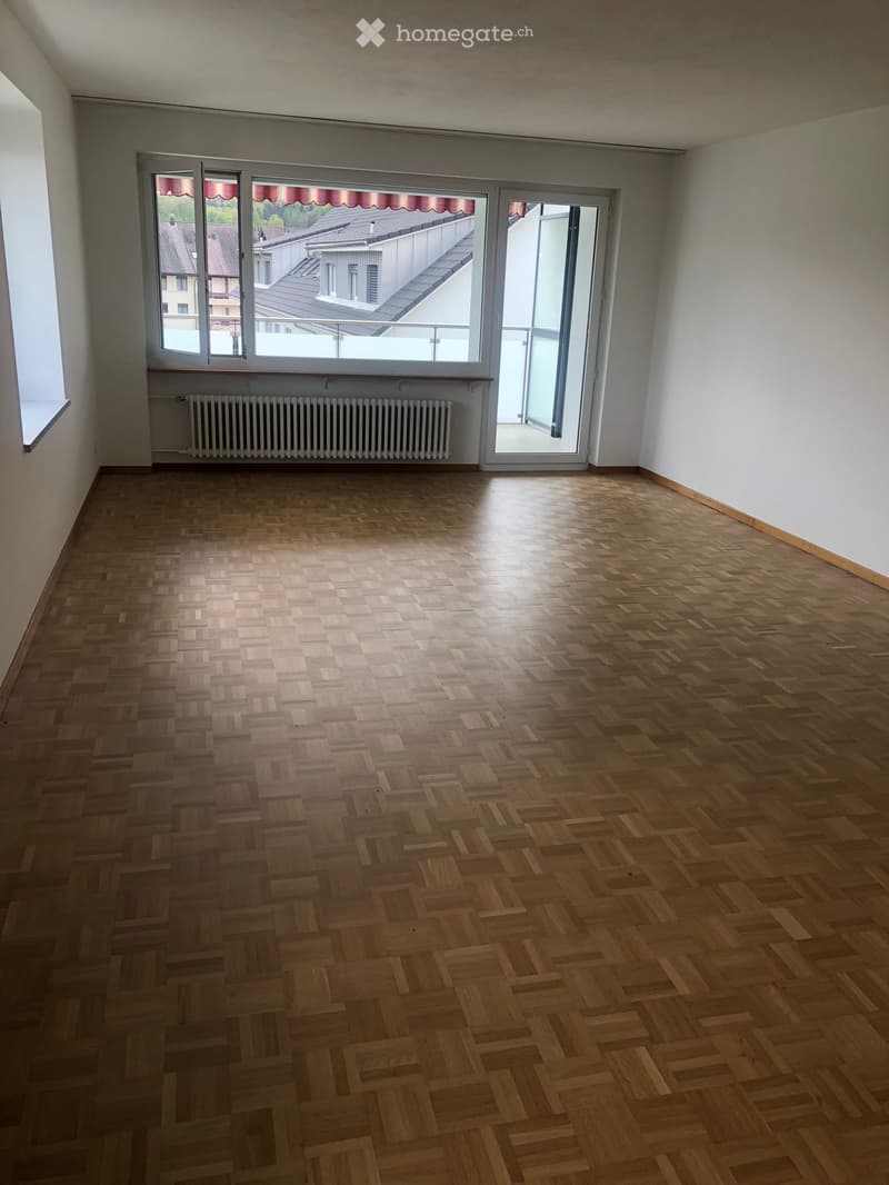 Empty room with wooden herringbone flooring, white walls, large windows, and a radiator. No furniture or equipment present.