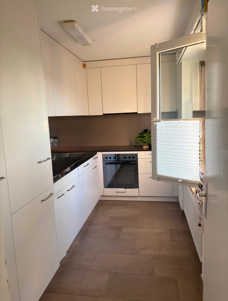 Modern kitchen with white cabinets, a built-in oven, and a radiator along the wall. The space features tiled flooring and an open window with blinds.