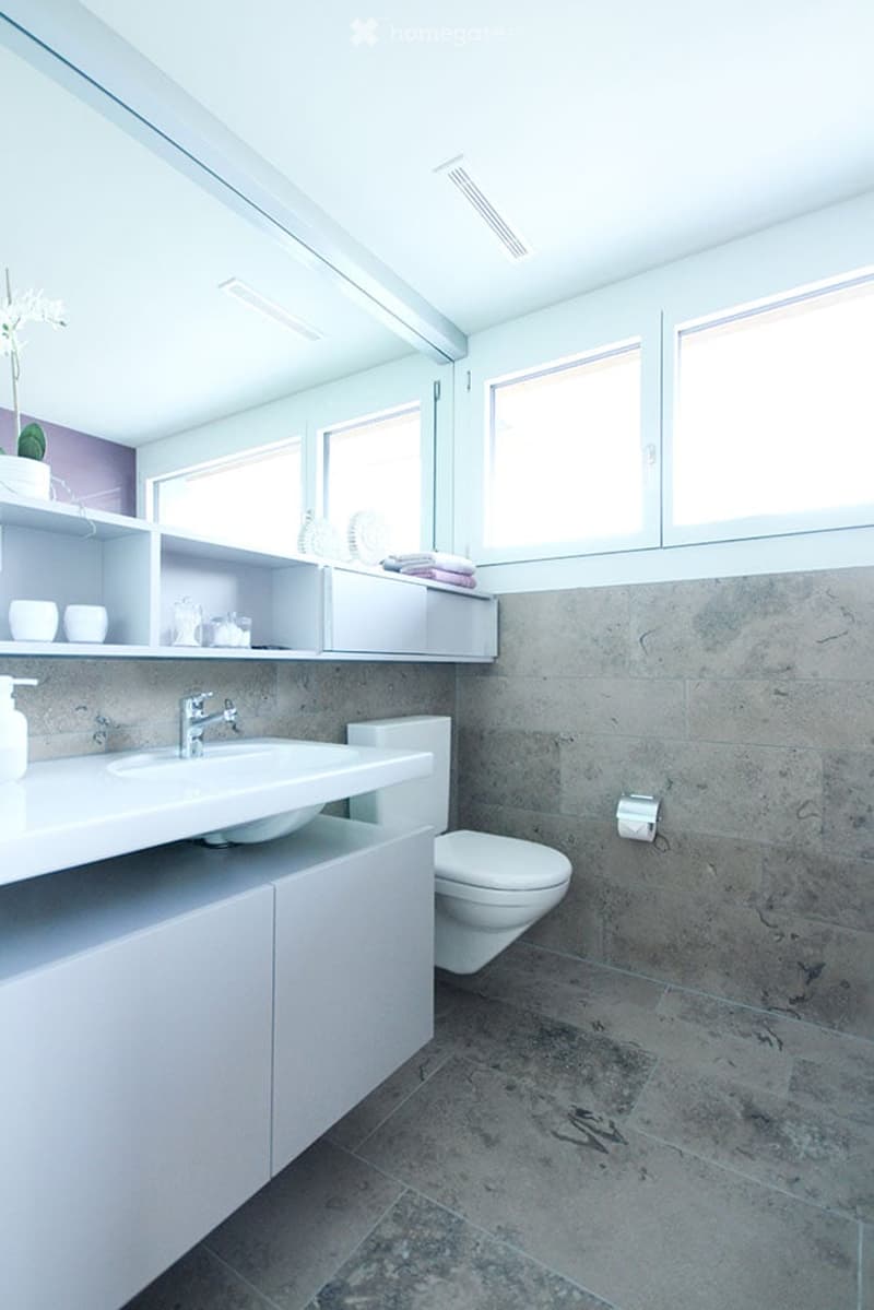 Modern bathroom with white cabinets, a sink, toilet, and shelves holding towels and decorative items. The walls are tiled in a neutral tone, and the space is well-lit with natural light from large windows.