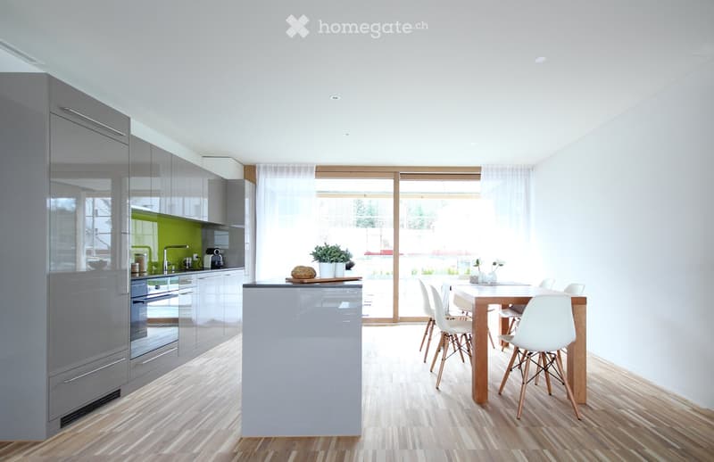 Modern kitchen with grey cabinets, a central island, a dining table with white chairs, and a view through large windows.