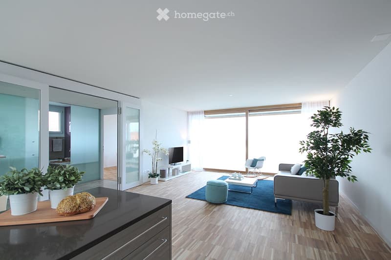 Bright living room with wooden flooring, large windows, grey sofa, coffee table, ottoman, armchair, television, and potted plants.