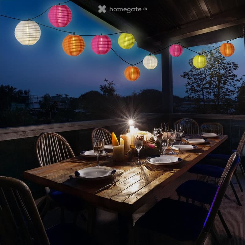 A wooden dining table set for a meal with plates, glasses, and candles, surrounded by colorful paper lanterns and blue chairs, set against a twilight backdrop with trees.