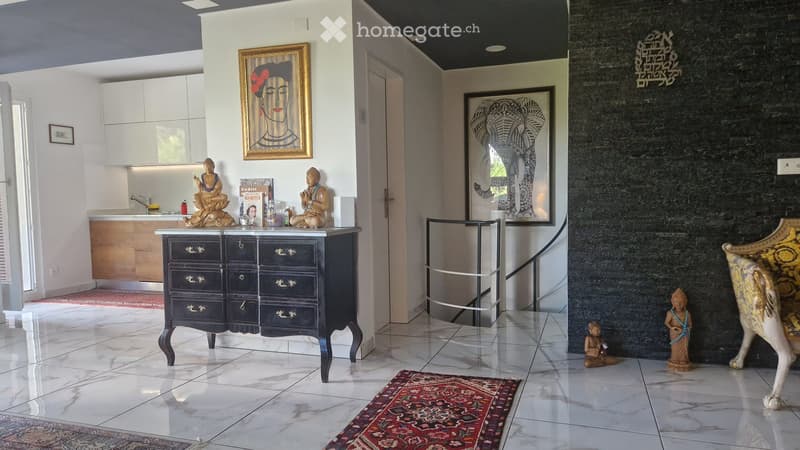 A modern, well-lit hallway with a polished tile floor, a black cabinet with gold handles, two decorative statues, a framed art piece, a kitchen area with a sink and cabinets, and a patterned rug on the floor.