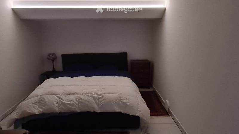 Small bedroom with a neatly made bed, a nightstand, and a lamp. The room has white walls and a built-in shelf above the bed.