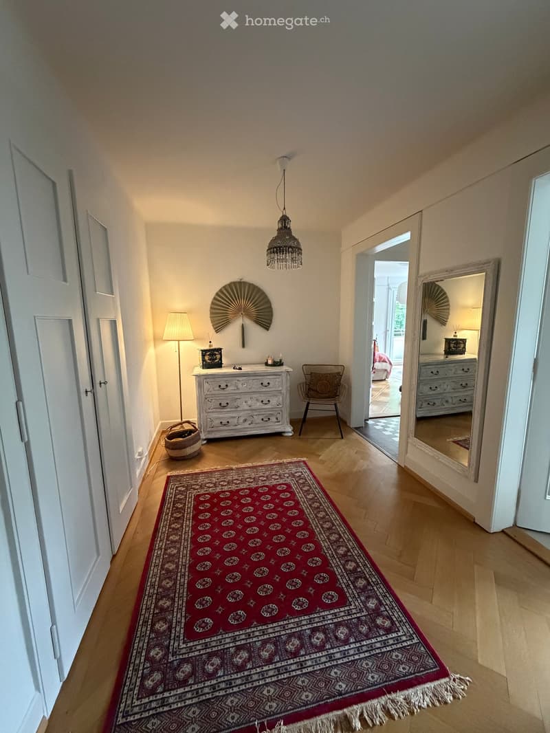 Hallway with wooden flooring, red patterned rug, white walls, and a chandelier hanging from the ceiling. There are doors on both sides leading to other rooms.
