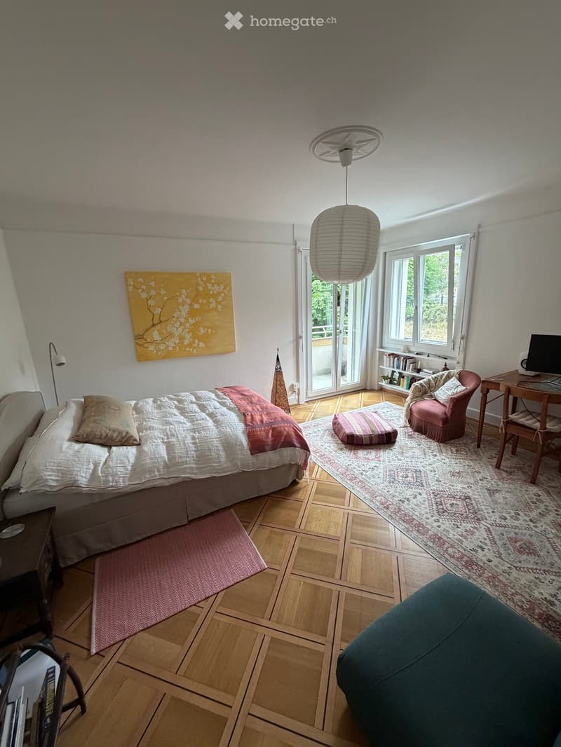 Cozy bedroom with a bed, sofa, chair, small table with laptop, bookshelf, and decorative rug. Features large windows and a sliding glass door leading to a balcony.