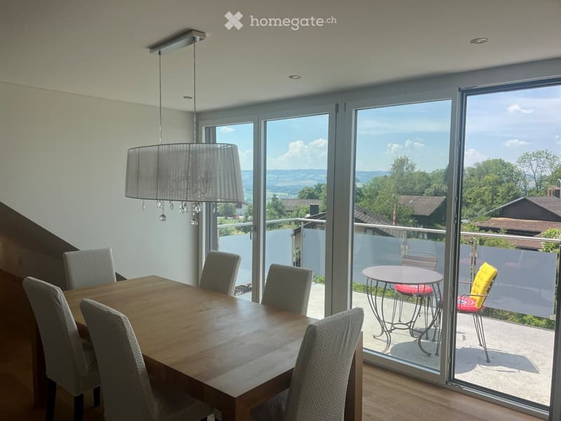 Bright dining area with a wooden table and chairs, large glass doors leading to a balcony with outdoor furniture and scenic countryside view.