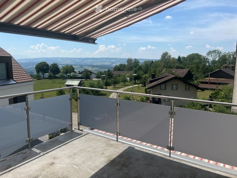 Balcony with metal railing, glass panels, and striped awning, overlooking a green landscape with houses and trees.