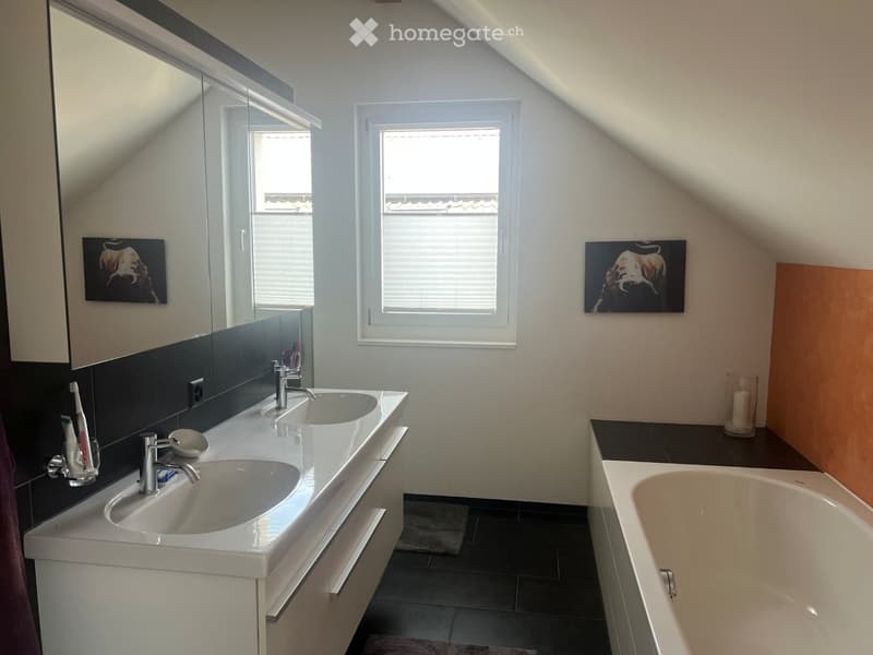 Modern bathroom with double sink, bathtub, large mirror, and window. Features include a sleek vanity with storage, a candle on the windowsill, and artwork on the walls.