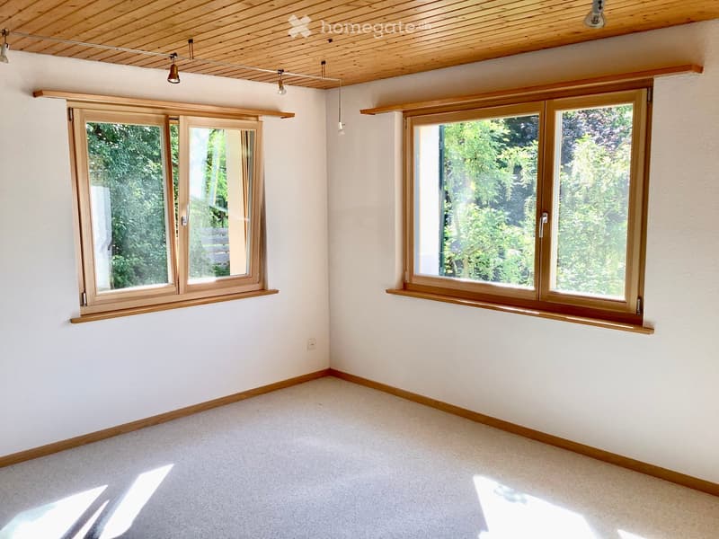 Empty room with light-colored walls, carpeted floor, and large wooden-framed windows with wooden sills. The room has a wooden ceiling with recessed lighting fixtures.