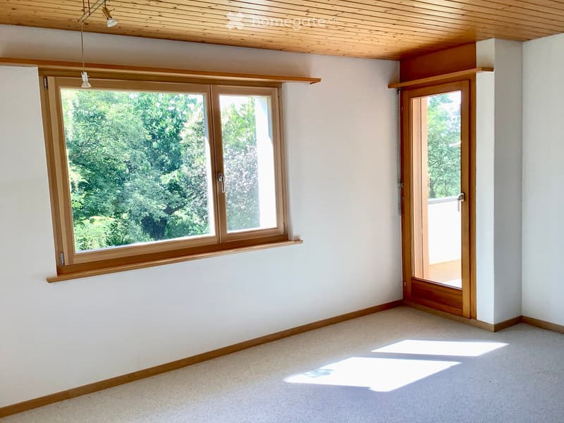 Empty room with white walls, wooden trim, large windows, and a sliding door leading outside.
