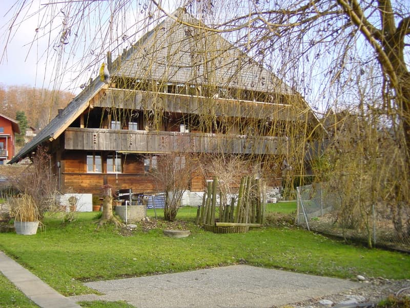 Two-story wooden house with a balcony, surrounded by a well-maintained garden with trees and shrubs.