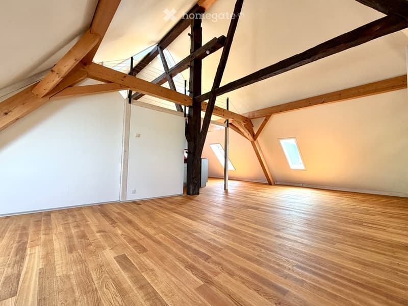 An empty attic space with wooden beams, wooden flooring, and skylight windows, featuring exposed wooden beams and white walls.