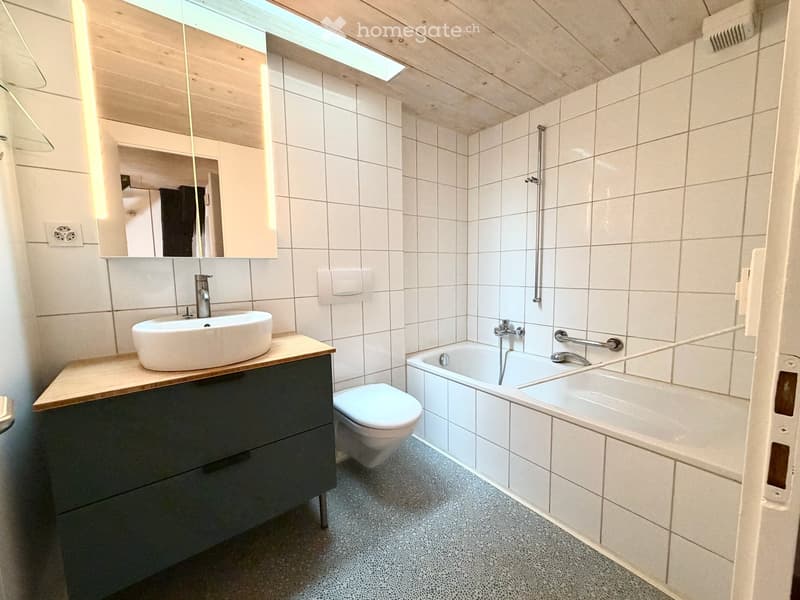 Modern bathroom with white tiles, a wooden countertop sink, a toilet, a bathtub, and a large mirror above the sink.