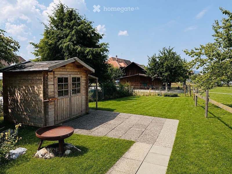 Well-maintained garden with a wooden shed, a metal fire pit, a concrete pathway, and a wooden fence. The area is surrounded by lush green grass and trees, with a larger house visible in the background.